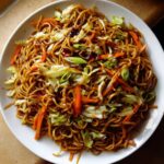 A close-up overhead shot of a white plate piled high with savory fried noodles mixed with shredded cabbage and carrots.