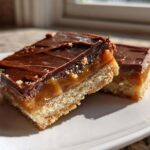 Two decadent, layered twix cookie bars featuring a shortbread base, thick caramel, and a rich chocolate topping, served on a white plate.