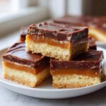 Three stacked squares of rich 3-Layer twix cookie bars showing shortbread, caramel, and chocolate layers.