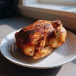 A perfectly cooked, crispy-skinned air fryer whole chicken resting on a white plate, illuminated by natural light.