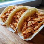 Close-up of three soft tortillas filled generously with saucy, shredded chicken tacos on a wooden cutting board.