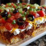 Cross-section view of a thick slice of 7 layer dip showing layers of crust, chili/bean mixture, sour cream, cheese, tomatoes, olives, and green onions.
