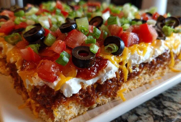 Cross-section view of a thick slice of 7 layer dip showing layers of crust, chili/bean mixture, sour cream, cheese, tomatoes, olives, and green onions.
