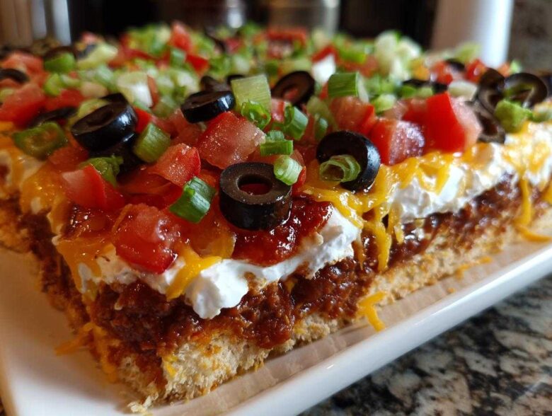 Cross-section view of a thick slice of 7 layer dip showing layers of crust, chili/bean mixture, sour cream, cheese, tomatoes, olives, and green onions.