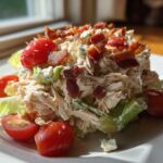A mound of creamy blt chicken salad topped with crispy bacon bits and fresh cherry tomatoes on a white plate.