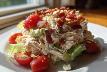 A mound of creamy blt chicken salad topped with crispy bacon bits and fresh cherry tomatoes on a white plate.