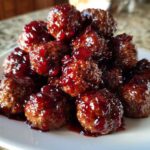A close-up of a pile of glazed cranberry meatballs glistening with a dark red sauce on a white plate.