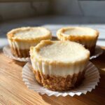 Close-up of three perfect mini cheesecake bites with graham cracker crusts sitting on a wooden cutting board.