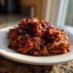 A mound of tender, saucy pulled chicken glistening under natural light on a white plate.