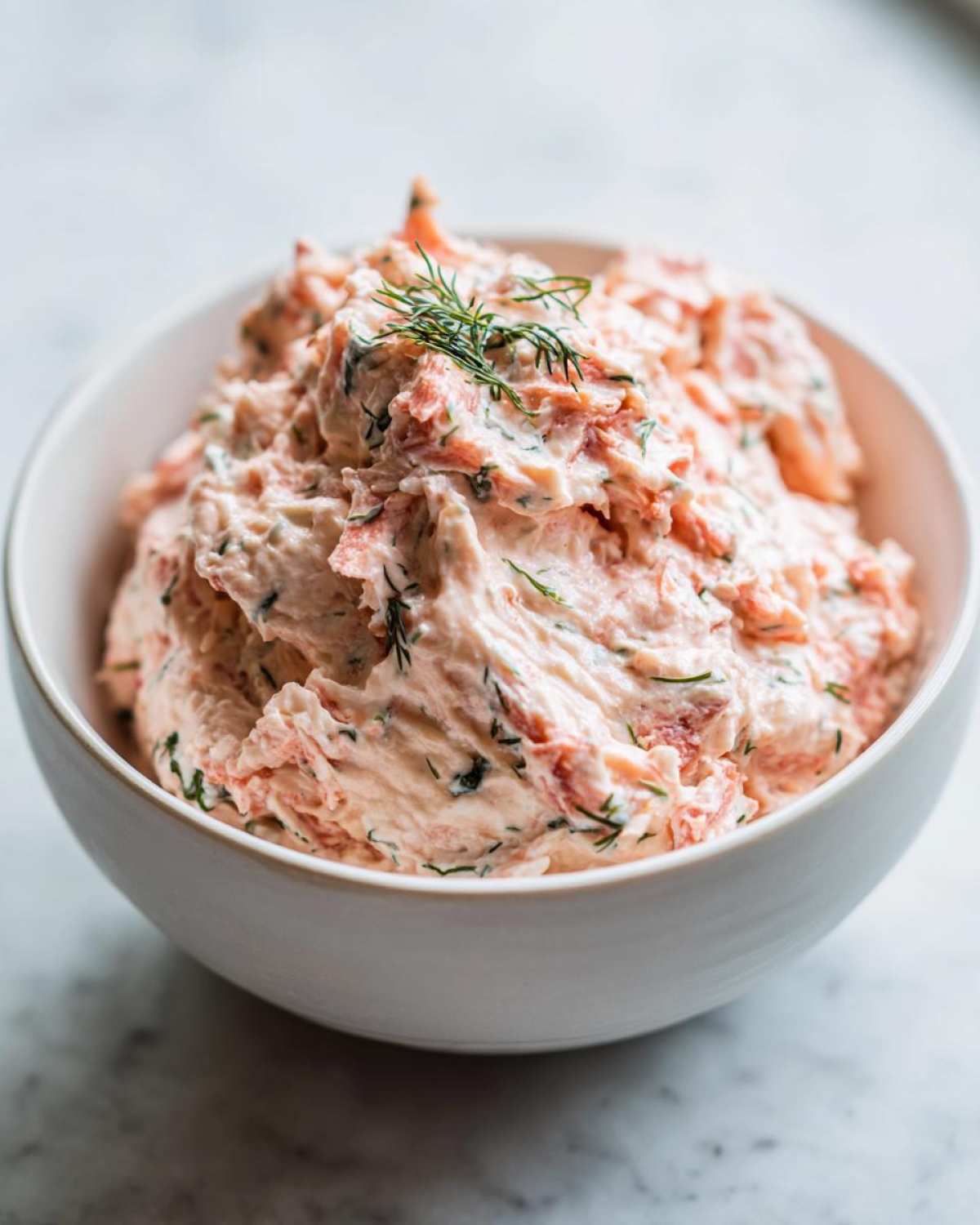 Close-up of creamy smoked salmon dip mixed with herbs, garnished with fresh dill, in a white bowl.