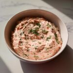 A white bowl filled with creamy, pink smoked salmon dip, garnished generously with fresh dill.