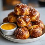 A stack of golden brown, salted soft pretzel bites served on a plate next to a small bowl of bright yellow cheese sauce.