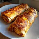 Two golden-brown baked chicken chimichangas with melted cheese, resting on a white plate.