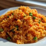 A close-up of fluffy, bright orange Spanish rice mixed with green peas and diced carrots served on a white plate.