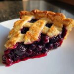 Close-up of a juicy slice of blueberry pie recipe featuring a golden lattice crust and rich, dark blueberry filling.