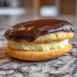 A close-up of a single boston cream pie cookies sandwich with vanilla cream filling and chocolate glaze.