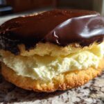 A single, decadent boston cream pie cookie featuring vanilla filling sandwiched between two cakes and topped with shiny chocolate ganache.