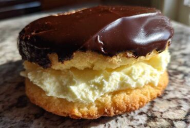 A single, decadent boston cream pie cookie featuring vanilla filling sandwiched between two cakes and topped with shiny chocolate ganache.