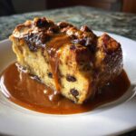 A close-up of a slice of bread pudding with bourbon sauce, featuring raisins, on a white plate.