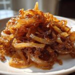 Close-up of a mound of deeply browned and glossy caramelized onions served on a white plate.