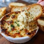 Hand dipping a toasted bread slice into a hot, cheesy, baked bread dip garnished with parsley.