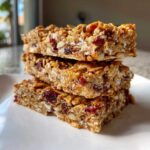 Three chewy homemade granola bars stacked on a white plate featuring oats, seeds, and dried cranberries.