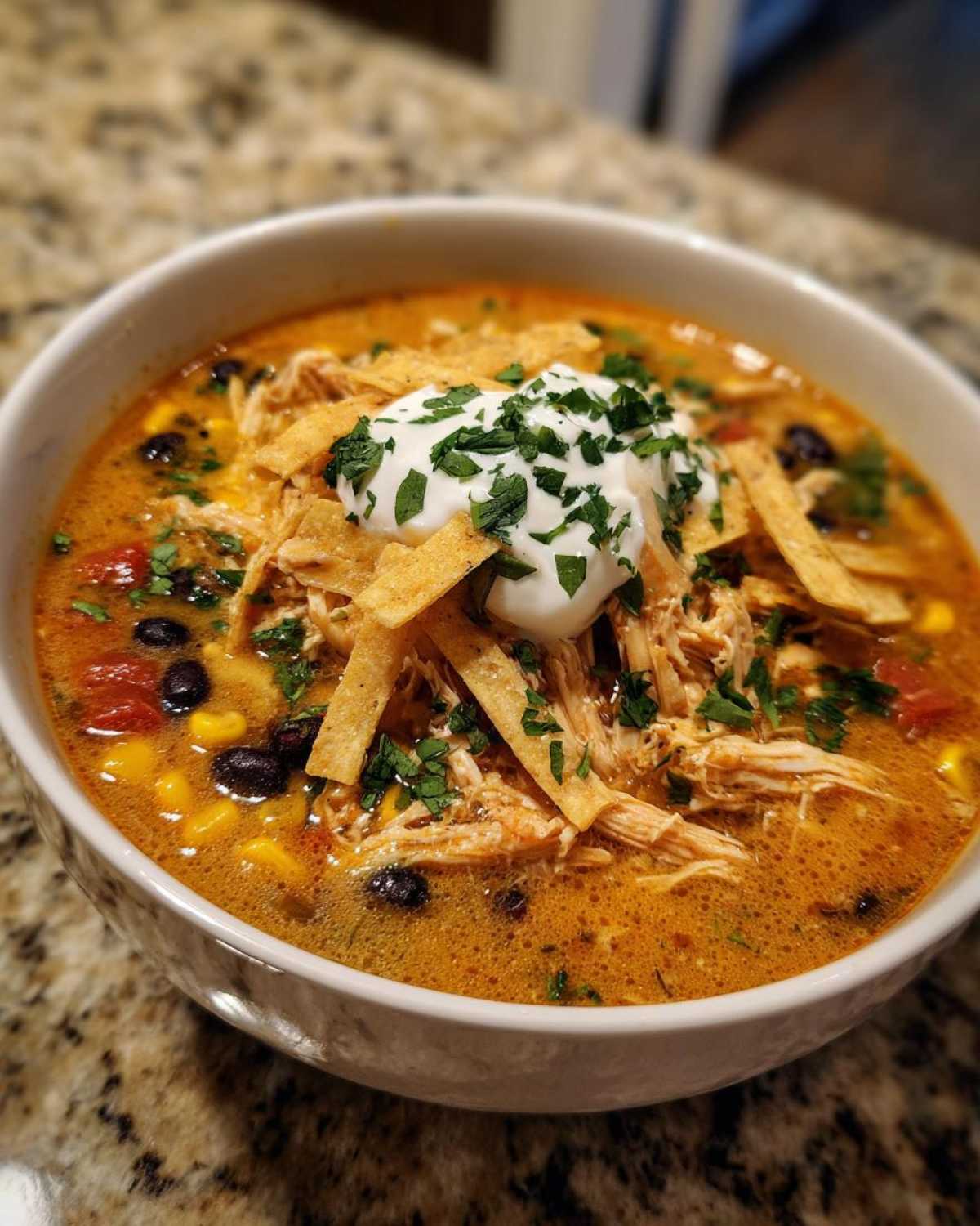 A close-up of a bowl of rich, orange-hued chicken tortilla soup topped with shredded chicken, black beans, corn, tortilla strips, and sour cream.