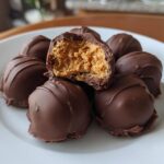 A pile of chocolate-covered peanut butter balls on a white plate, one is bitten open showing the creamy filling.