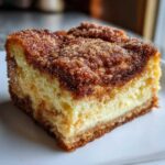 A close-up shot of a thick slice of churro cheesecake on a white plate, showing the creamy filling and cinnamon sugar topping.
