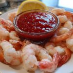 A plate of chilled, cooked shrimp arranged around a small bowl of bright red cocktail sauce, garnished with a lemon wedge for a perfect shrimp cocktail.