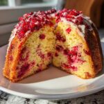 A slice of moist cranberry orange cake, showing bright red cranberries embedded in the yellow crumb, topped with white glaze and chopped cranberries.