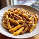 A close-up shot of a bowl filled with creamy beef pasta (penne shape) coated in a rich, cheesy sauce with ground beef.