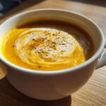 Close-up of bright orange cauliflower soup in a white mug, swirled with cream and topped with fresh black pepper.