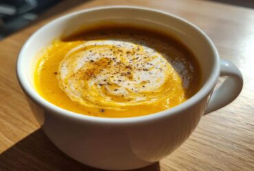 Close-up of bright orange cauliflower soup in a white mug, swirled with cream and topped with fresh black pepper.