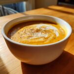 Close-up of a bowl of rich, orange-hued cauliflower soup topped with a white cream swirl and cracked black pepper.