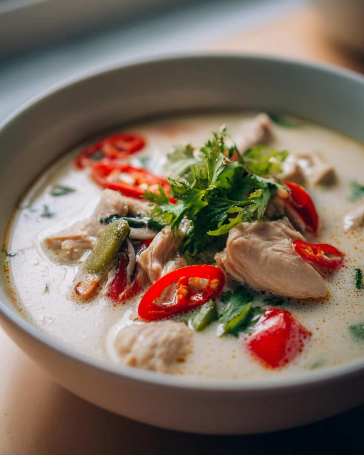 Close-up of a creamy, white coconut soup filled with chunks of chicken, bright red chili slices, and fresh cilantro.