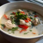 Close-up of a white bowl filled with creamy chicken coconut soup, garnished with fresh cilantro and red chili slices.