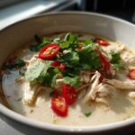 Close-up of a bowl of creamy chicken coconut soup topped with shredded chicken, fresh cilantro, and bright red chili slices.