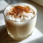 Close-up of a chilled glass of creamy coquito topped with frothy foam and a dusting of cinnamon.