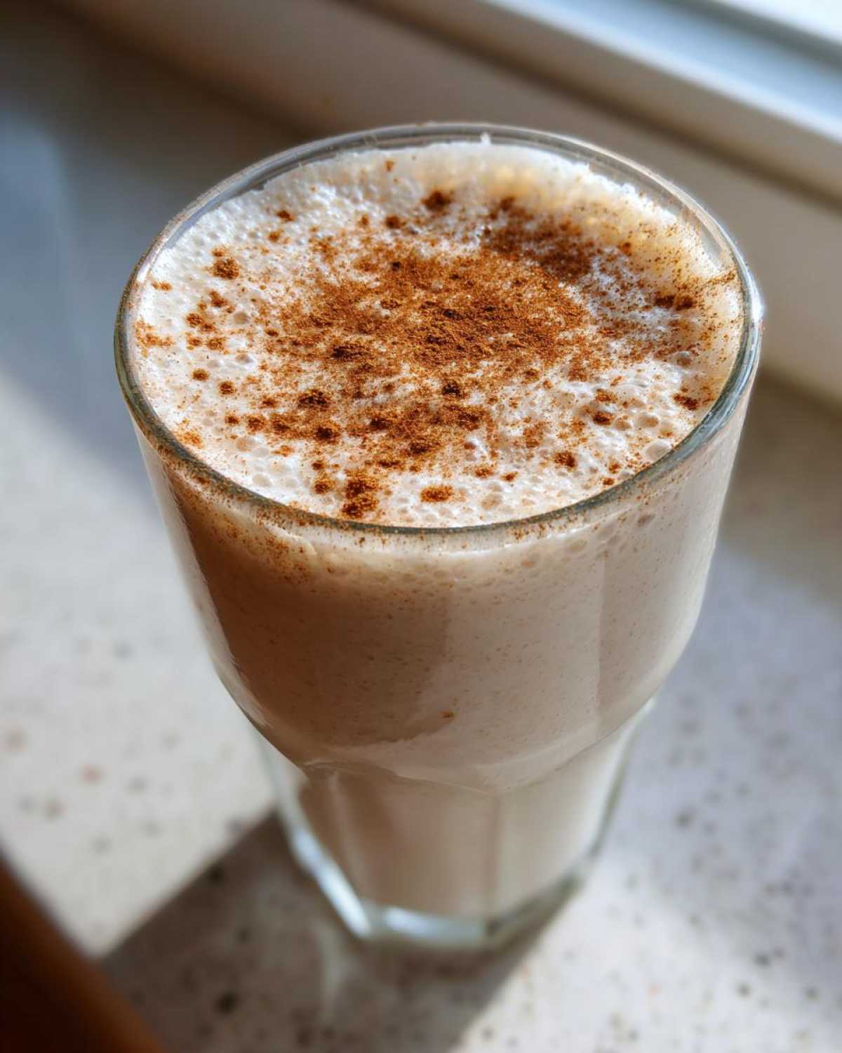 A tall glass filled with creamy, frothy coquito recipe, generously dusted with ground cinnamon.
