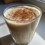 Close-up of a glass filled with creamy coquito recipe, topped with a generous dusting of ground cinnamon.