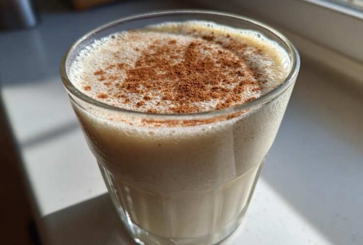 Close-up of a glass filled with creamy coquito recipe, topped with a generous dusting of ground cinnamon.