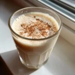 A creamy glass of coquito recipe drink, topped with a dusting of ground cinnamon, sitting near a window.