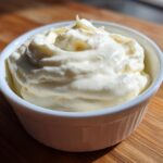 A close-up of thick, creamy homemade alfredo sauce swirled high in a small white ramekin on a wooden surface.