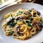 Close-up of creamy mushroom pasta featuring fettuccine noodles, sliced brown mushrooms, and wilted spinach.