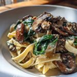 Close-up of creamy mushroom pasta featuring fettuccine noodles, sautéed mushrooms, and wilted spinach in a white bowl.