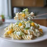 A spoonful of creamy pickle pasta salad lifted above a plate, showing rotini pasta, chopped pickles, and dill.