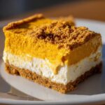A thick slice of pumpkin cheesecake bars showing a graham cracker crust, a white cream cheese layer, and a bright orange pumpkin layer topped with crumbs.