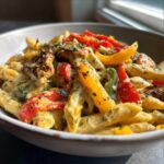 Close-up of creamy rasta pasta featuring penne, colorful bell peppers, and seasoned chicken pieces, garnished with herbs.