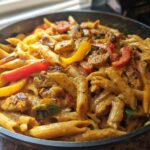 Close-up of creamy Rasta pasta mixed with seasoned chicken pieces and colorful bell peppers in a dark skillet.